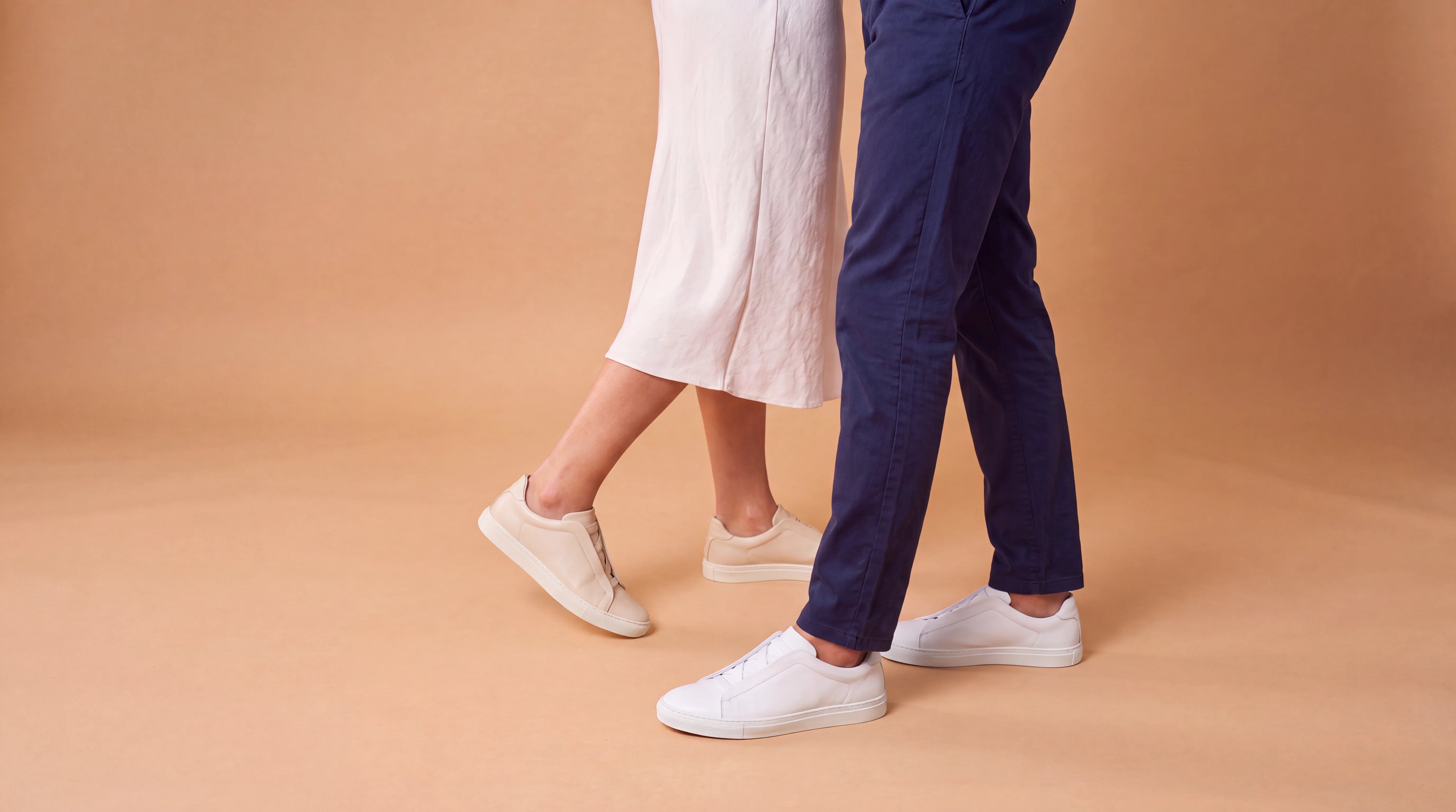 Two people walking side by side, shown from the waist down, wearing Aliverti white low-top laceless sneakers. One person wears a white midi dress and the other wears navy blue trousers, set against a solid warm tan studio backdrop.